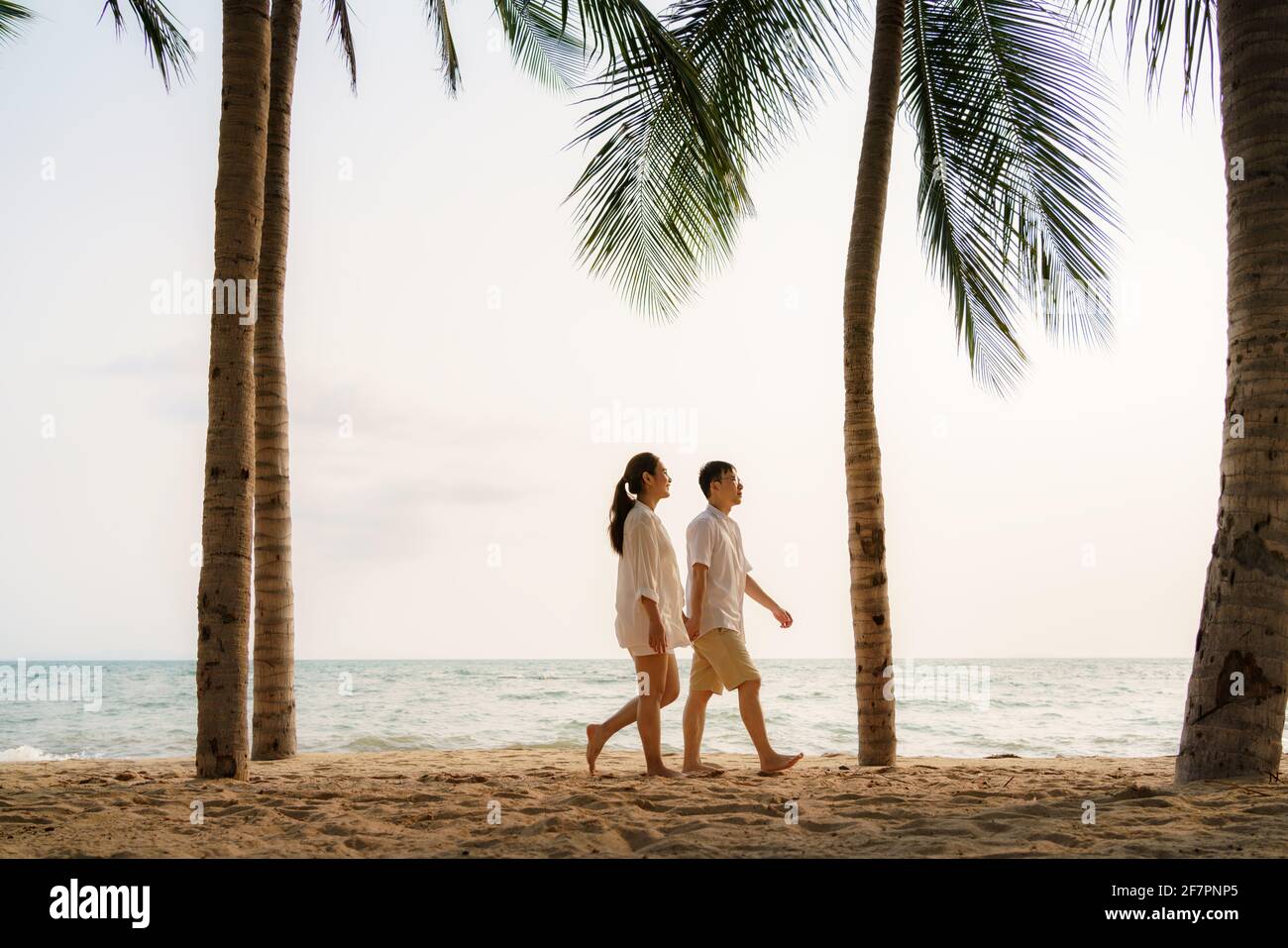 Asian couple is walking and holding hands on a beachfront beach sea with coconut trees while on vacation in the summer in Thailand. Stock Photo