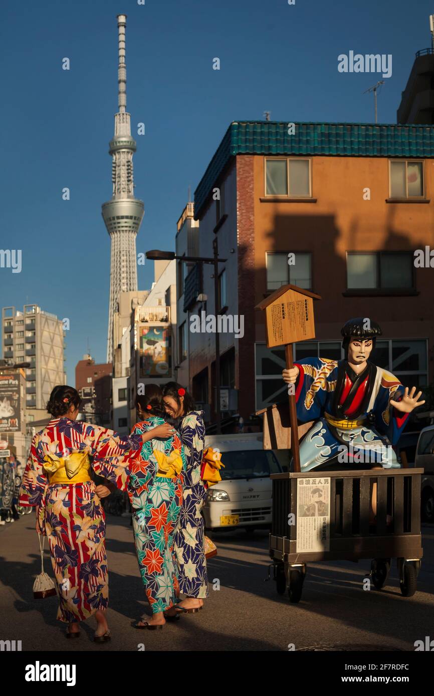 Three young women, wearing traditional yukatas, passing by an advertising Japanese character with Tokyo Sky Tree in the background, Asakusa, Tokyo Stock Photo