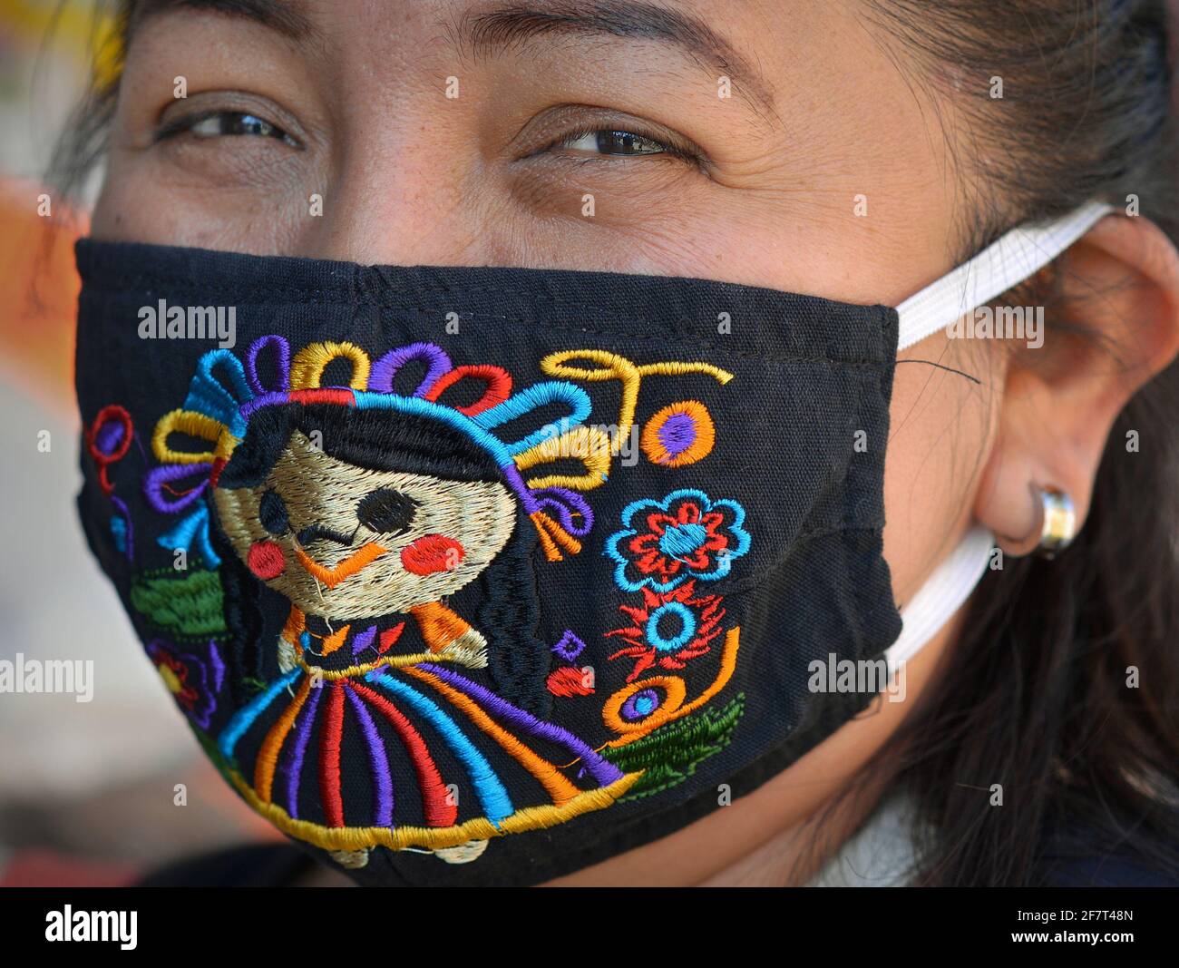 Mexican woman with smiling eyes wears a colorful washable embroidered cloth face mask and looks at the camera, during the global coronavirus pandemic. Stock Photo