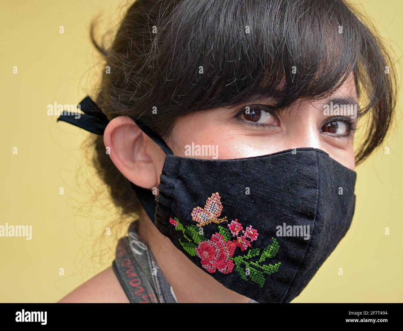 Young Mexican woman with beautiful dark eyes wears a cross-stitched embroidered cloth face mask with floral design during the coronavirus pandemic. Stock Photo