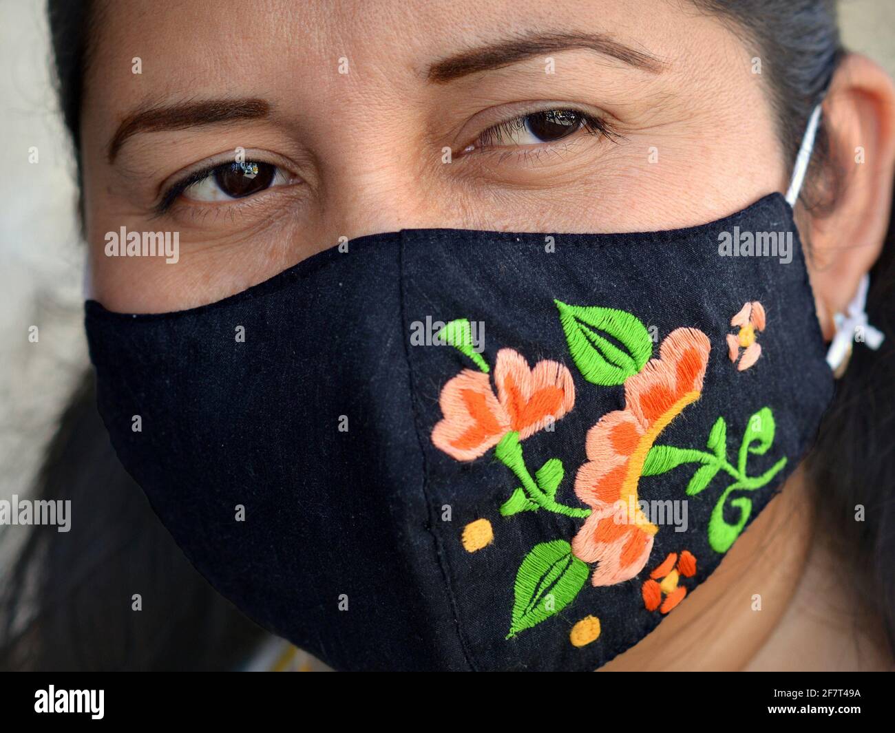 Young Mexican woman wears a colorful embroidered folkloric cloth face mask during the global coronavirus pandemic and smiles with her brown eyes. Stock Photo