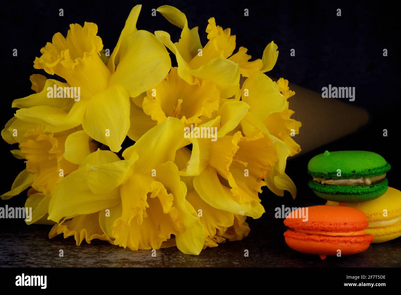 A bouquet of Daffodils in a paper cone and three colorful macarons (green, orange, yellow) are lying beside it. Stock Photo
