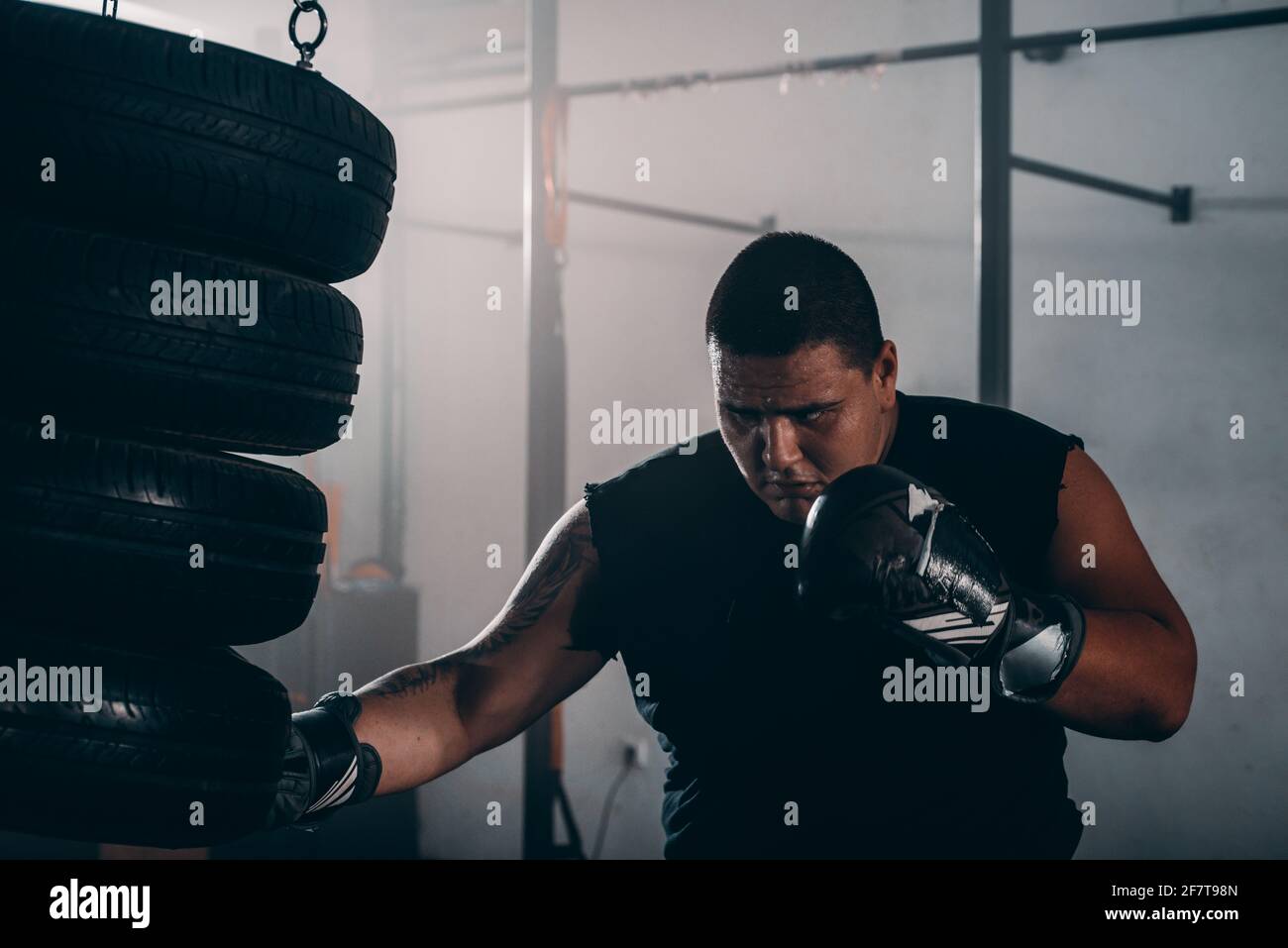 Handsome muscular man in boxing gloves is practicing with a punching tires while warming up
