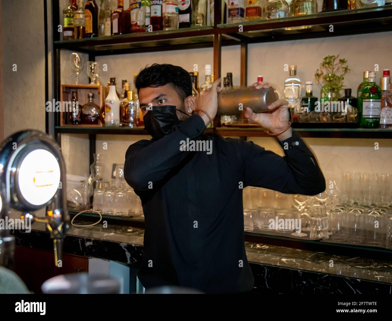 DUBAI, UNITED ARAB EMIRATES - Mar 25, 2021: bartender shaking a cocktail with a mask on in black with alcohol on the shelve at the back Stock Photo
