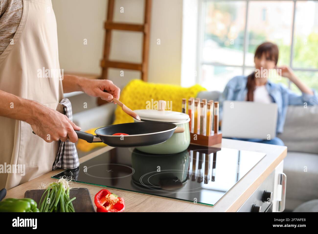 Young man cooking dinner while his wife is using laptop in kitchen Stock Photo
