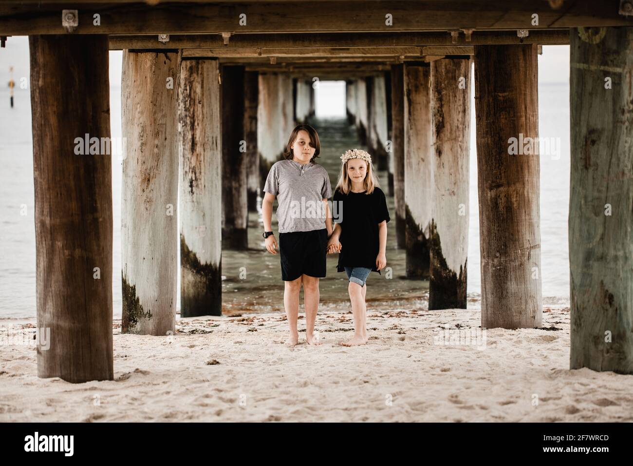 Caucasian girl standing underneath a pier at the beach wearing a flower head band holding hands with a boy Stock Photo