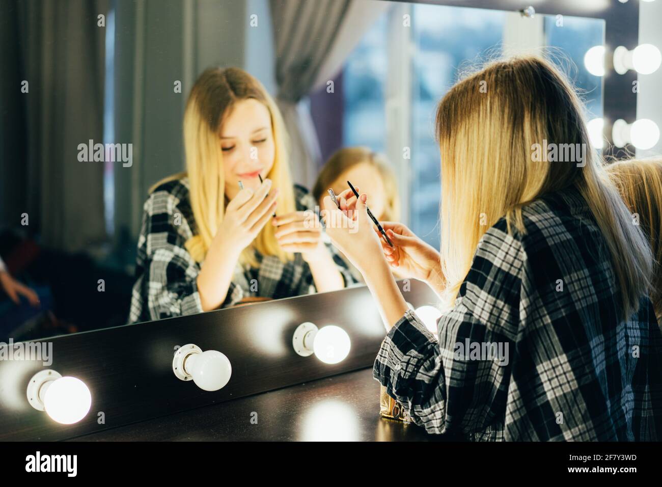 Teen in the dressing room in front of the mirror makes a make-up. Stylish girl in a plaid shirt with cosmetics. Stock Photo