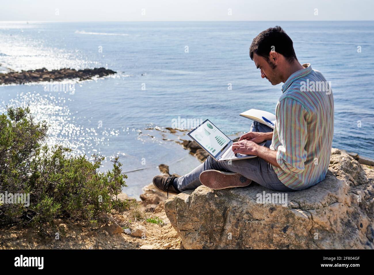 A young man uses his laptop and notepad while working remotely with the sea in the background Stock Photo