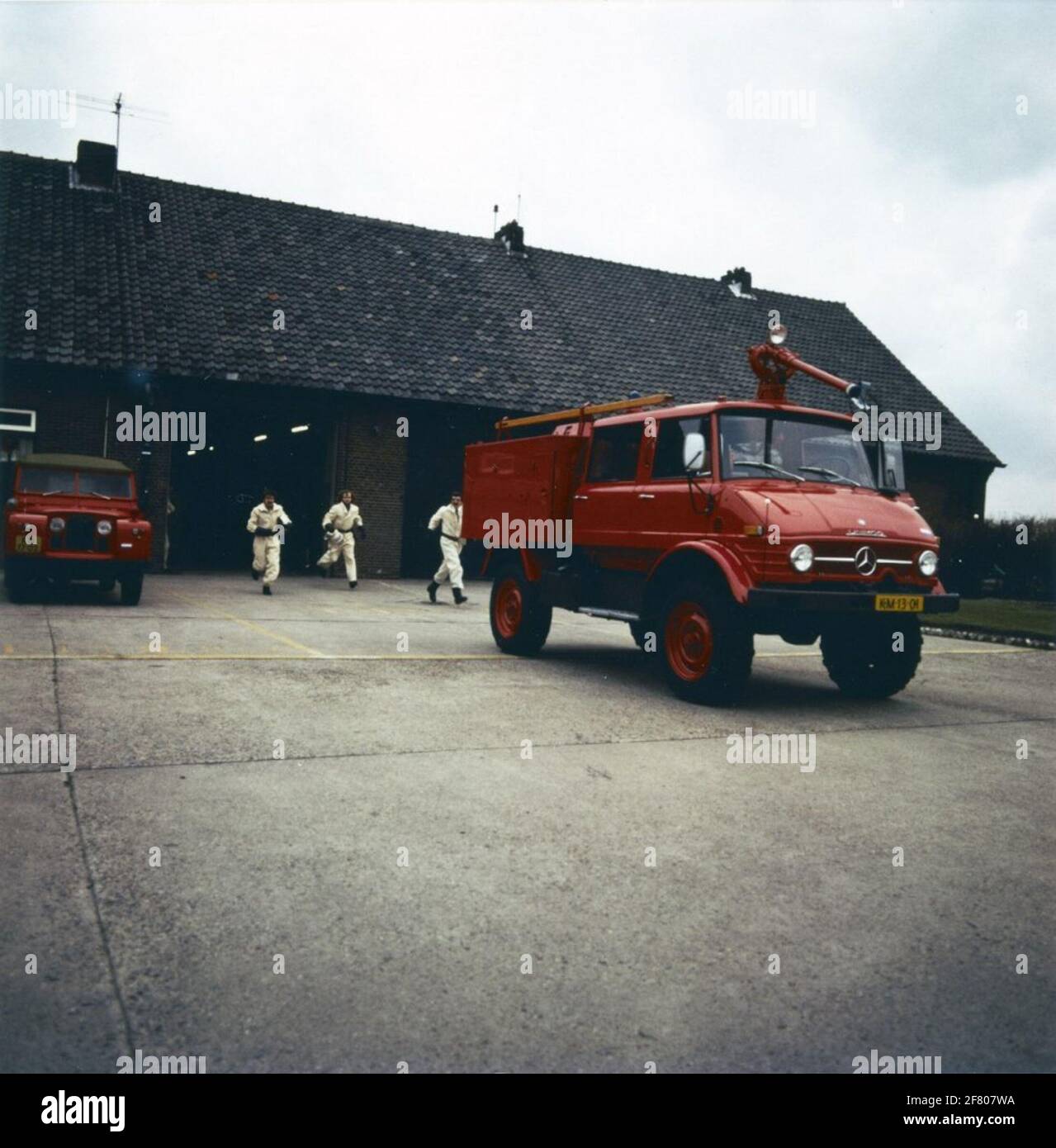 Firefighters at MarineLiegkamp Valkenburg run to a ready Mercedes-Benz Unimog in fire brigade. On the left is a Land Rover parked, also in fire brigade. Stock Photo