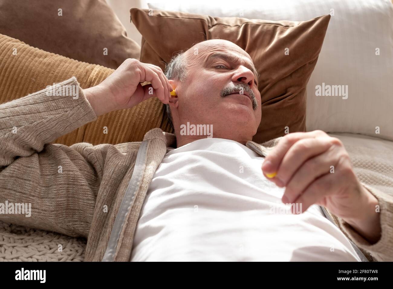 Caucasian senior stressed man holding a yellow earplug trying to sleep having insomnia resting at home alone Stock Photo