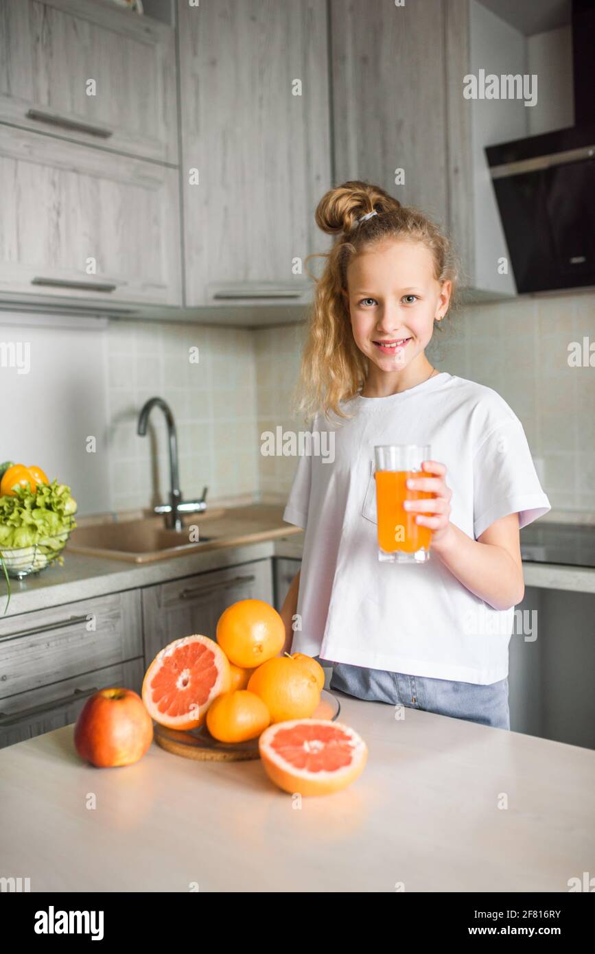 Caucasian child girl drinking orange juice indoors sitting at kitchen table. Kid healthy food nutrition concept. Stock Photo