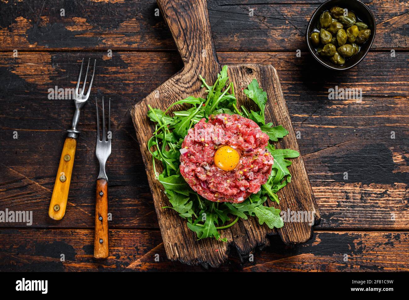 Tartar beef with a quail egg and arugula served on a cutting board. Dark wooden background. Top view Stock Photo