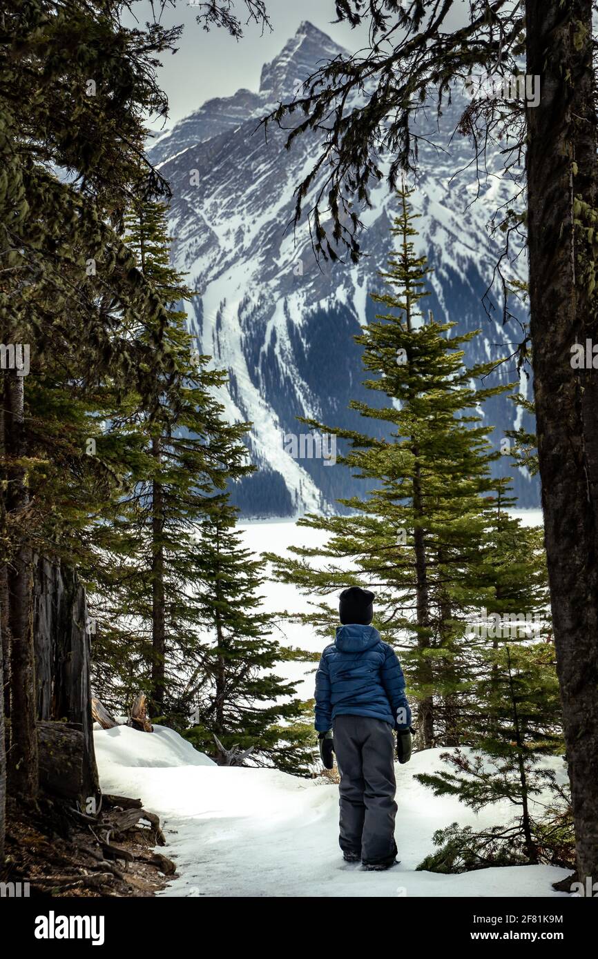 A young boy hiking on a snow covered mountain trail overlooking a frozen lake in the Canadian Rocky Mountains Stock Photo