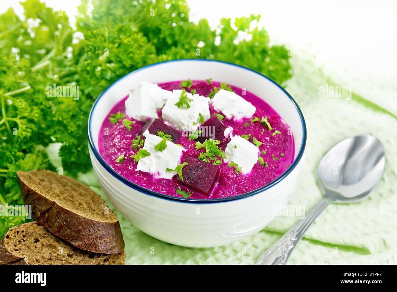 Soup puree of beetroot, potato and cream with salted cream cheese in a bowl on kitchen towel, spoon, bread and parsley on wooden board background Stock Photo
