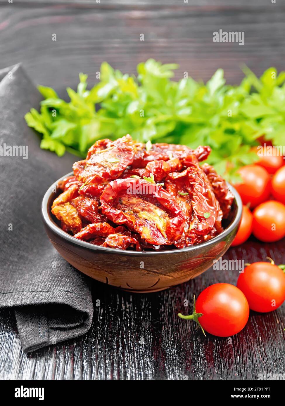 Sun-dried tomatoes in oil with thyme and basil in a bowl, napkin, fresh small tomatoes and parsley on black wooden board background Stock Photo