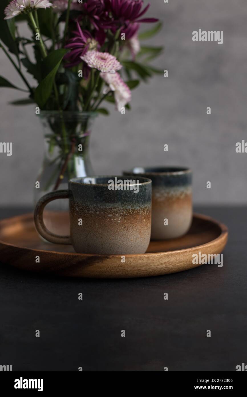 Two elegant rustic coffee cups on a wooden tray next to a blurred bouquet of dahlias. Stock Photo