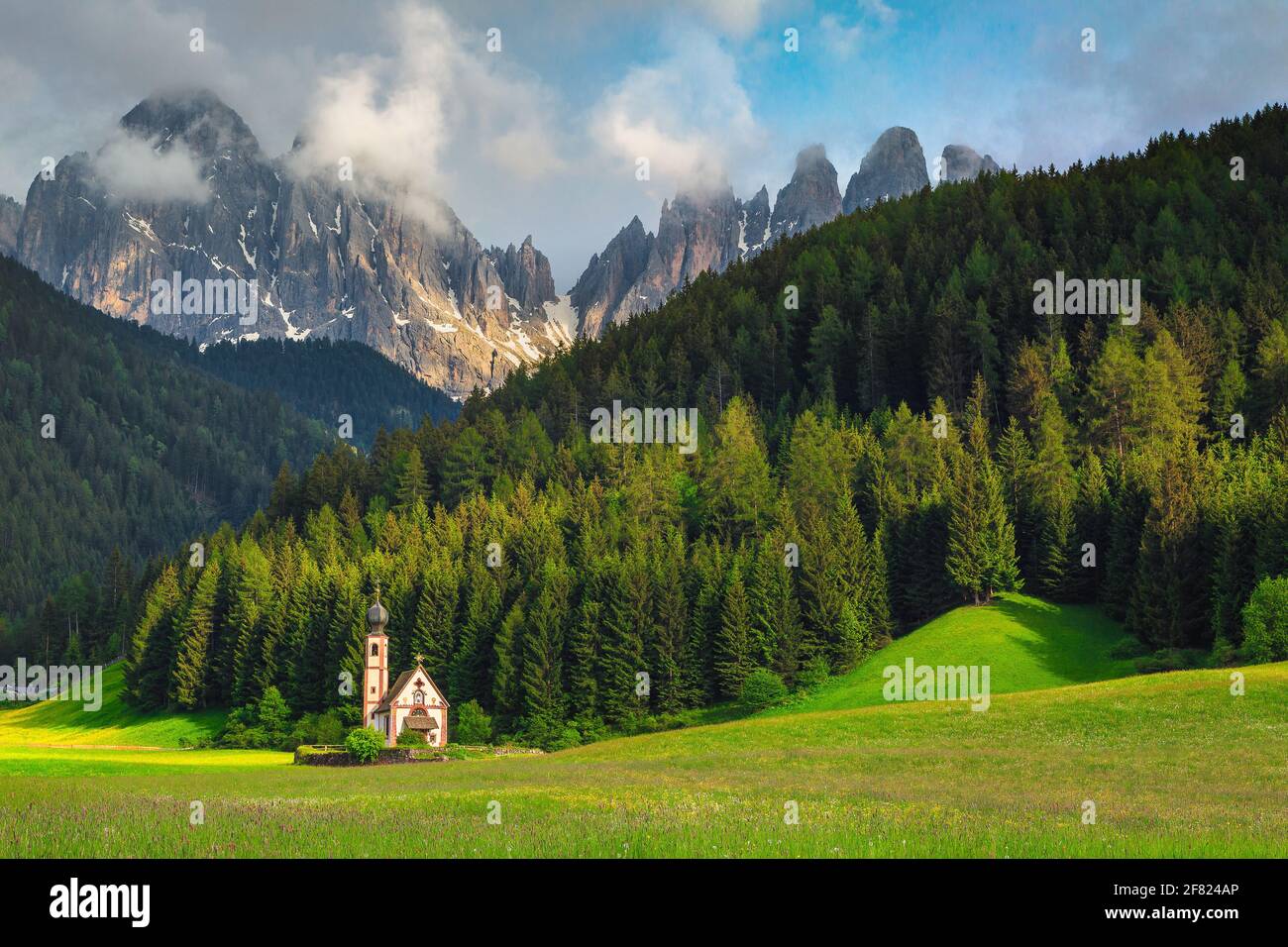 Great travel location, cute alpine St Johann church in Val di Funes valley with green fields and snowy mountains, Santa Maddalena village, Dolomites, Stock Photo