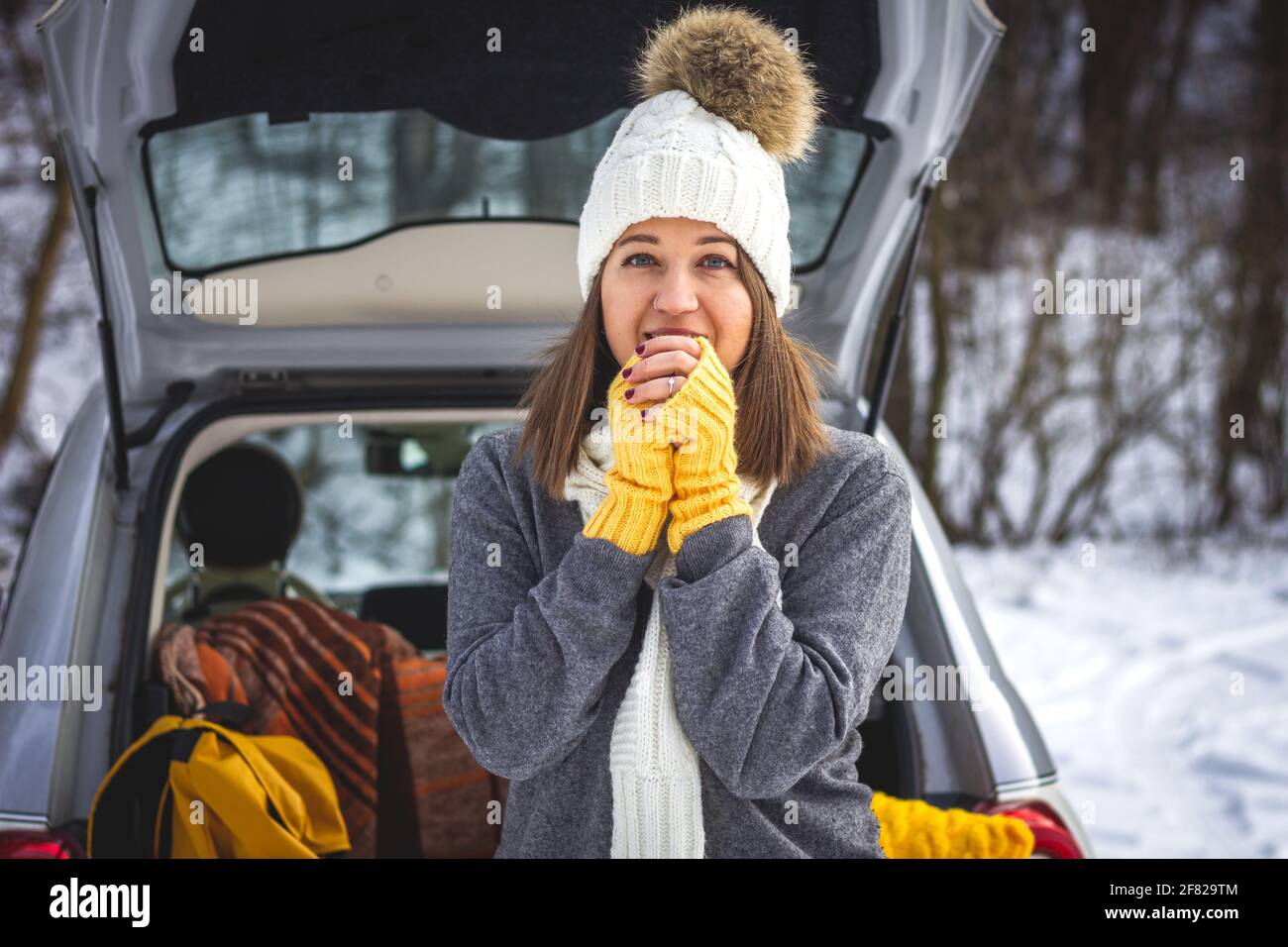 Young woman with knit hat warming up herself next to car during travel to winter vacation. Caucasian woman standing at outdoor parking lot. Road trip Stock Photo