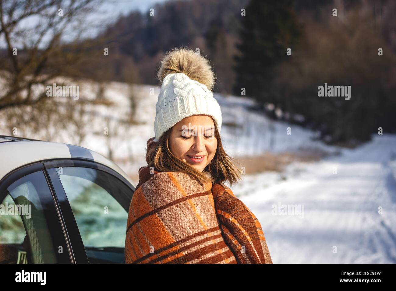 Young woman warming up herself in blanket on sunlight and resting next to car during winter vacation. Caucasian smiling woman with eyes closed Stock Photo