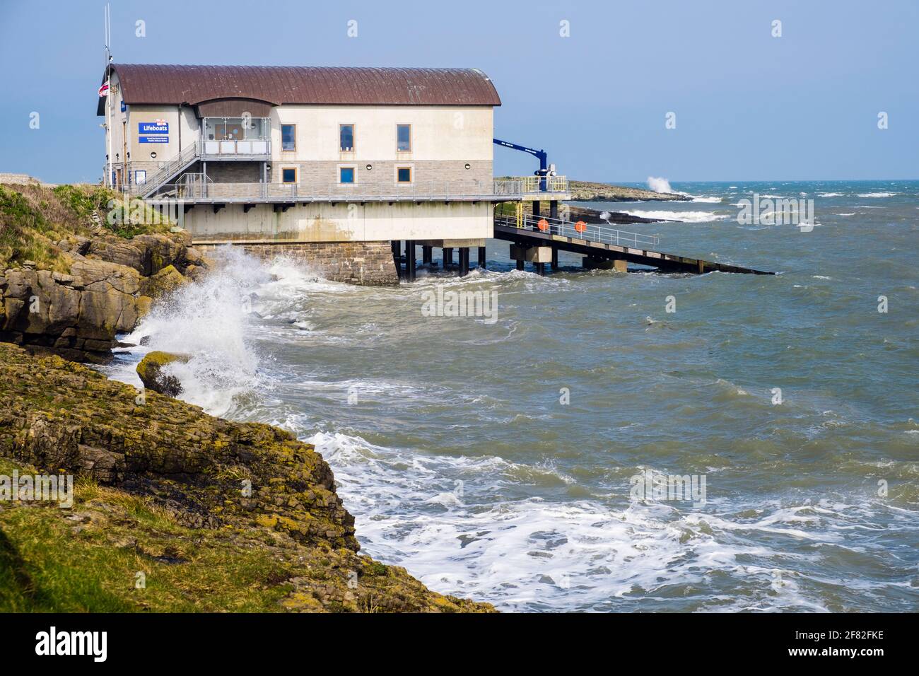 Rough choppy seas crashing on rocks at high tide by new RNLI lifeboat station boathouse in Moelfre, Isle of Anglesey, north Wales, UK, Britain Stock Photo