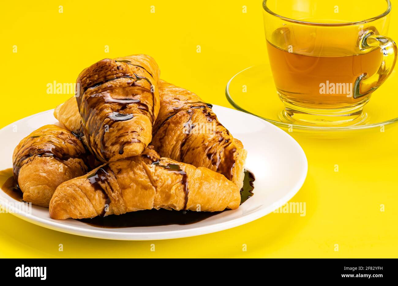 View pile of homemade croissants poured with chocolate topping in white ceramic dish and a cup of hot tea on yellow background with clipping path. Stock Photo