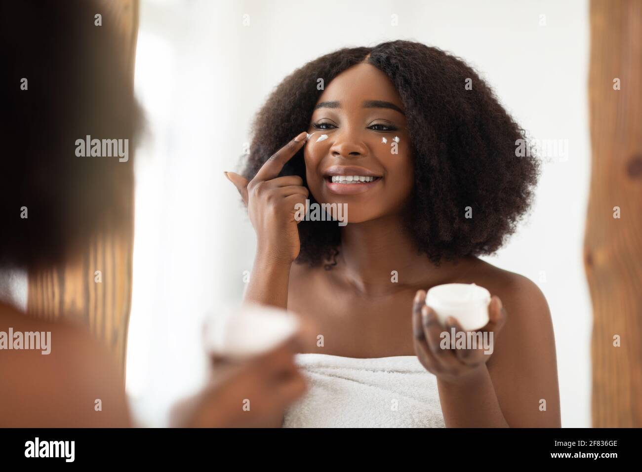 Young black woman in towel applying face cream after shower, looking in mirror, using natural cosmetic product at home Stock Photo