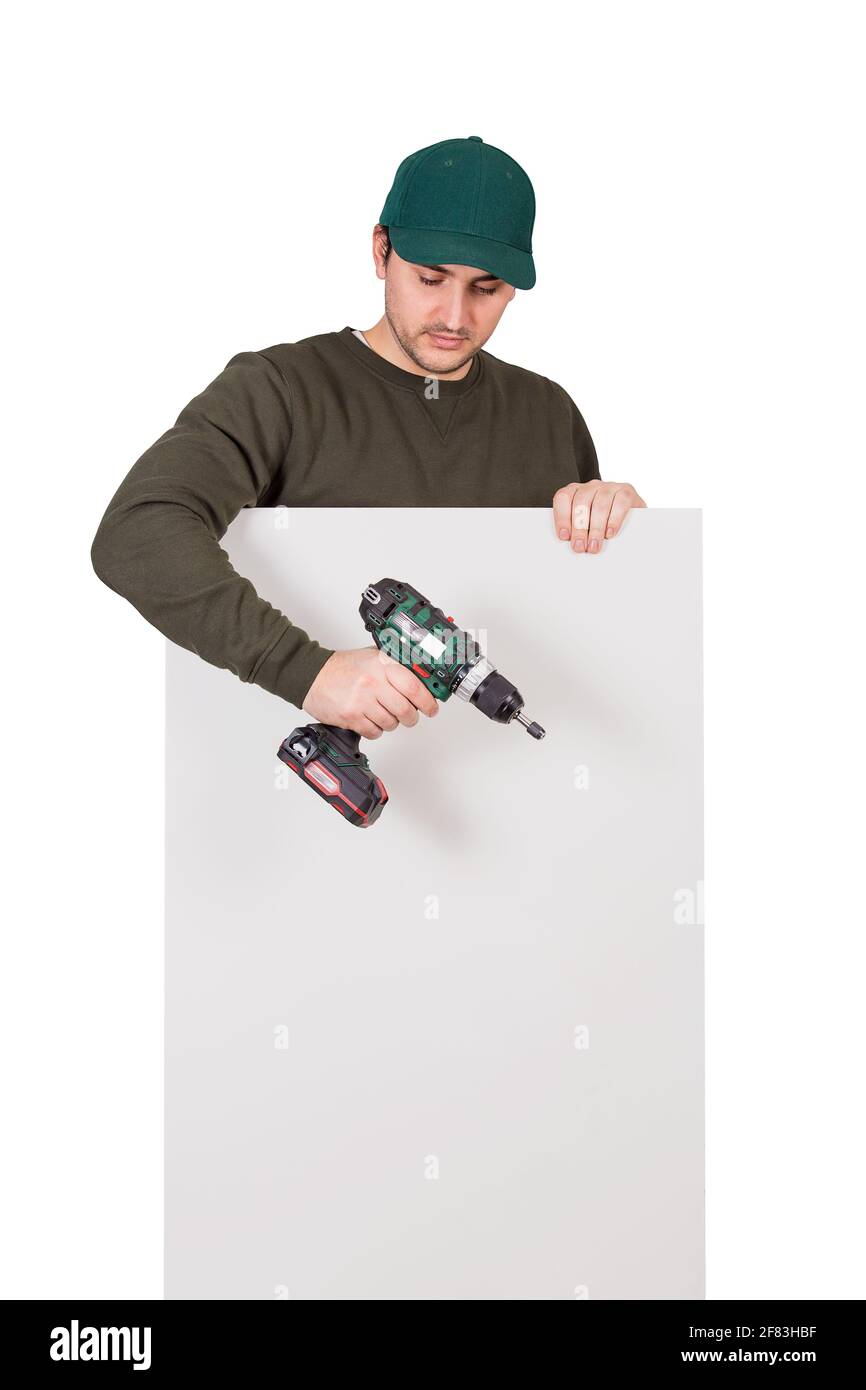Young man with a screwdriver or electric drill in his hand, stands behind a white panel. Renovation worker before installing interior finish plate or Stock Photo