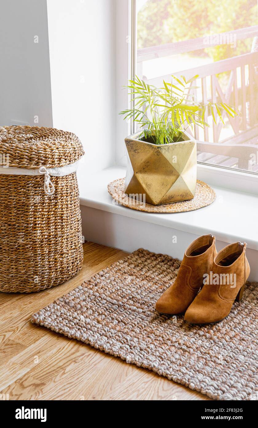 Hardwood floor with jute doormat, shoes and flower pot and seagrass laundry basket by window. Natural material objects in home concept. Home interior. Stock Photo