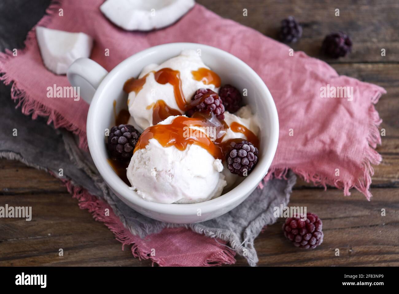 Coconut vegan ice cream in a white cup. Sundae balls and caramel. Ice cream with berries and raspberries. Wooden background, copy space. Stock Photo