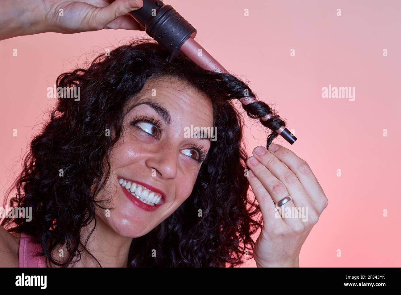 Young smiling woman with black curly hair uses tweezers or curling iron to get curls and waves in her hair. care and beauty concept. copy space. Stock Photo