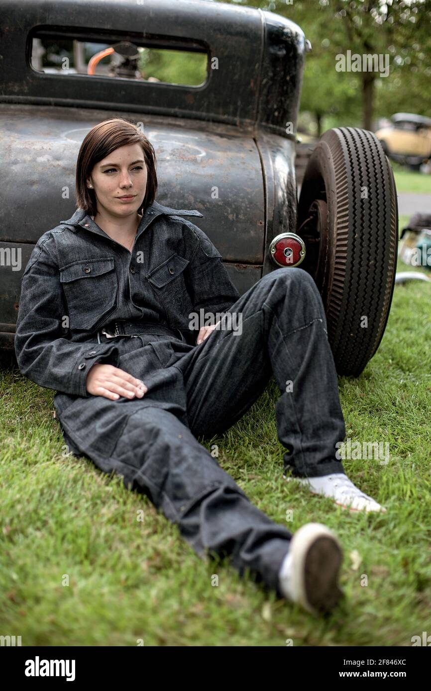 young beautiful woman in denim overall leaning on parked  hot rod car , Stock Photo