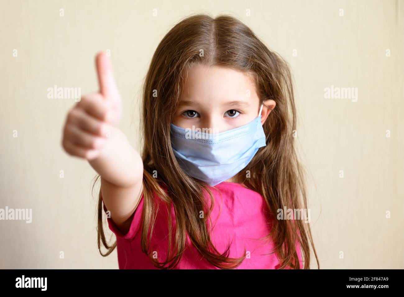 Kid in face mask for protection to corona virus shows thumb up, portrait of pretty little girl wearing medical mask due to COVID-19. Cute child stayin Stock Photo