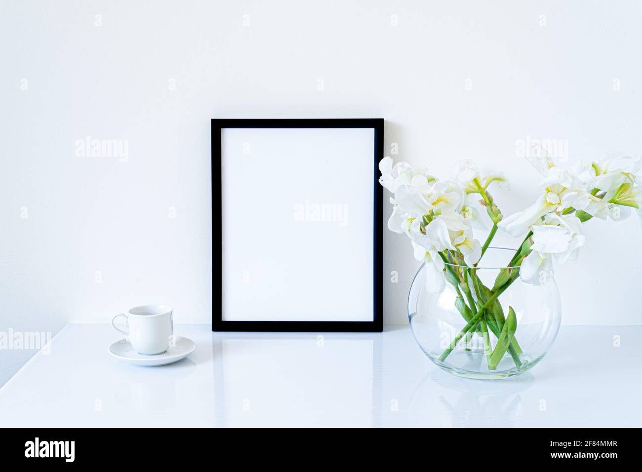Blank black frame mockup. Fresh flowers of white irises in glass vase in the shape of a sphere on a white table and coffee cup. White background, mini Stock Photo