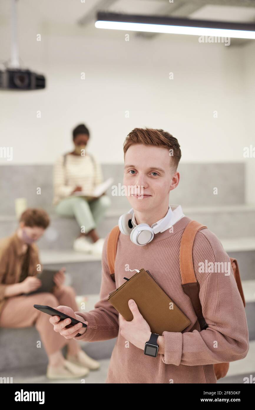 Vertical waist up portrait of young male student smiling at camera while standing in modern school lounge Stock Photo