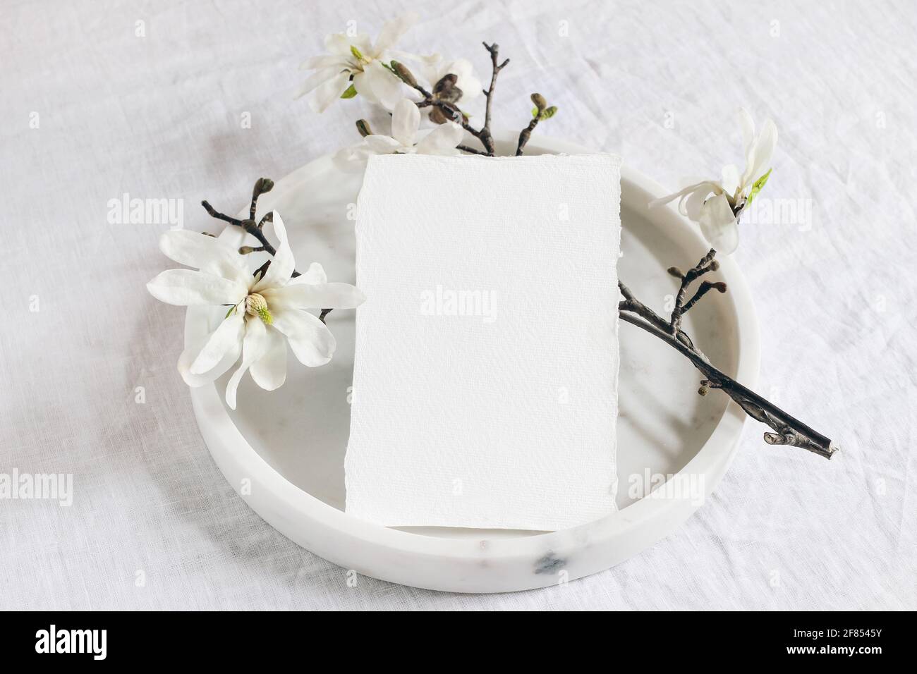 Wedding stationery mock-up scene. Blank vertical greeting card on marble tray. White linen tablecloth background with blooming magnolia stellata tree Stock Photo