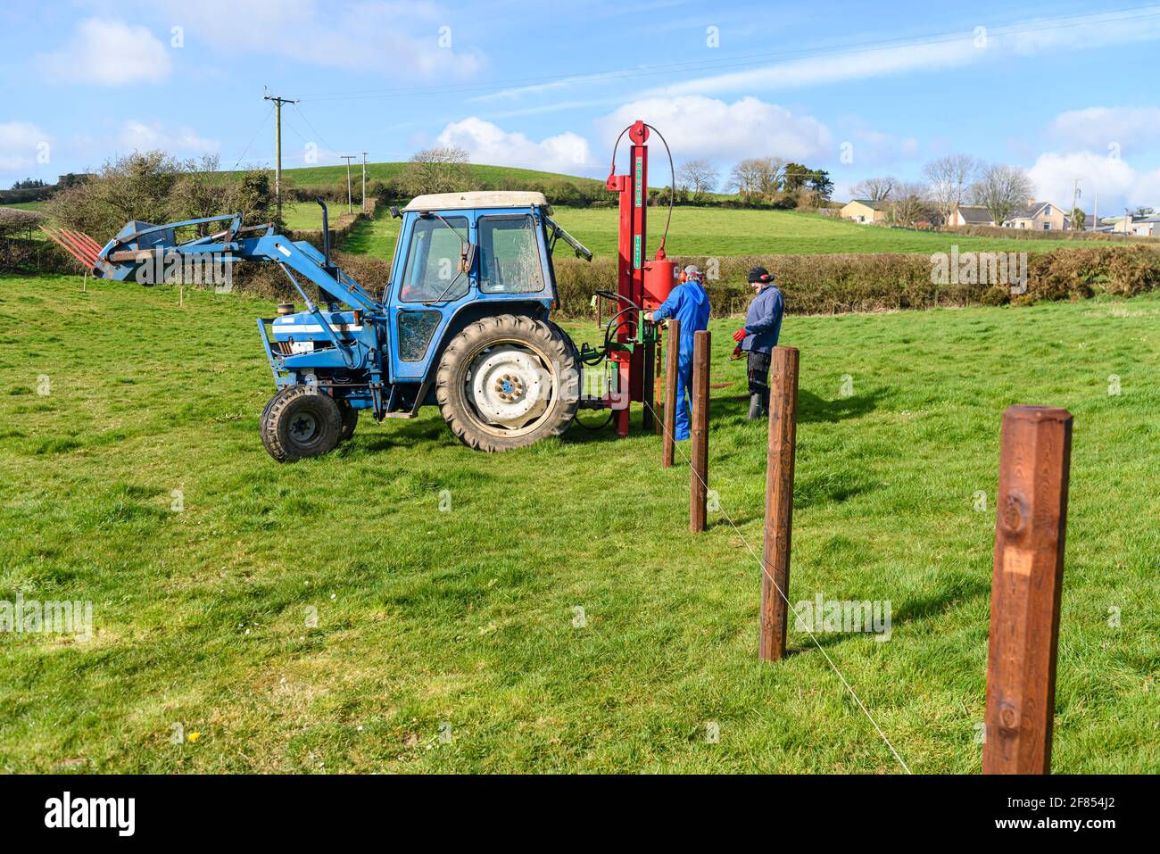 Two farmers men use a hydraulic post driver to install fence posts in a