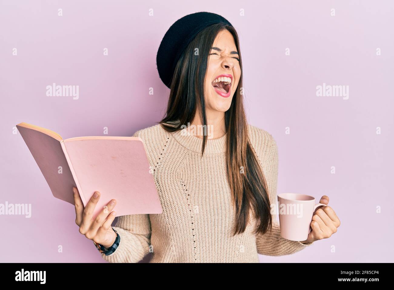 Young brunette woman reading a book and drinking a cup of coffee angry and mad screaming frustrated and furious, shouting with anger looking up. Stock Photo