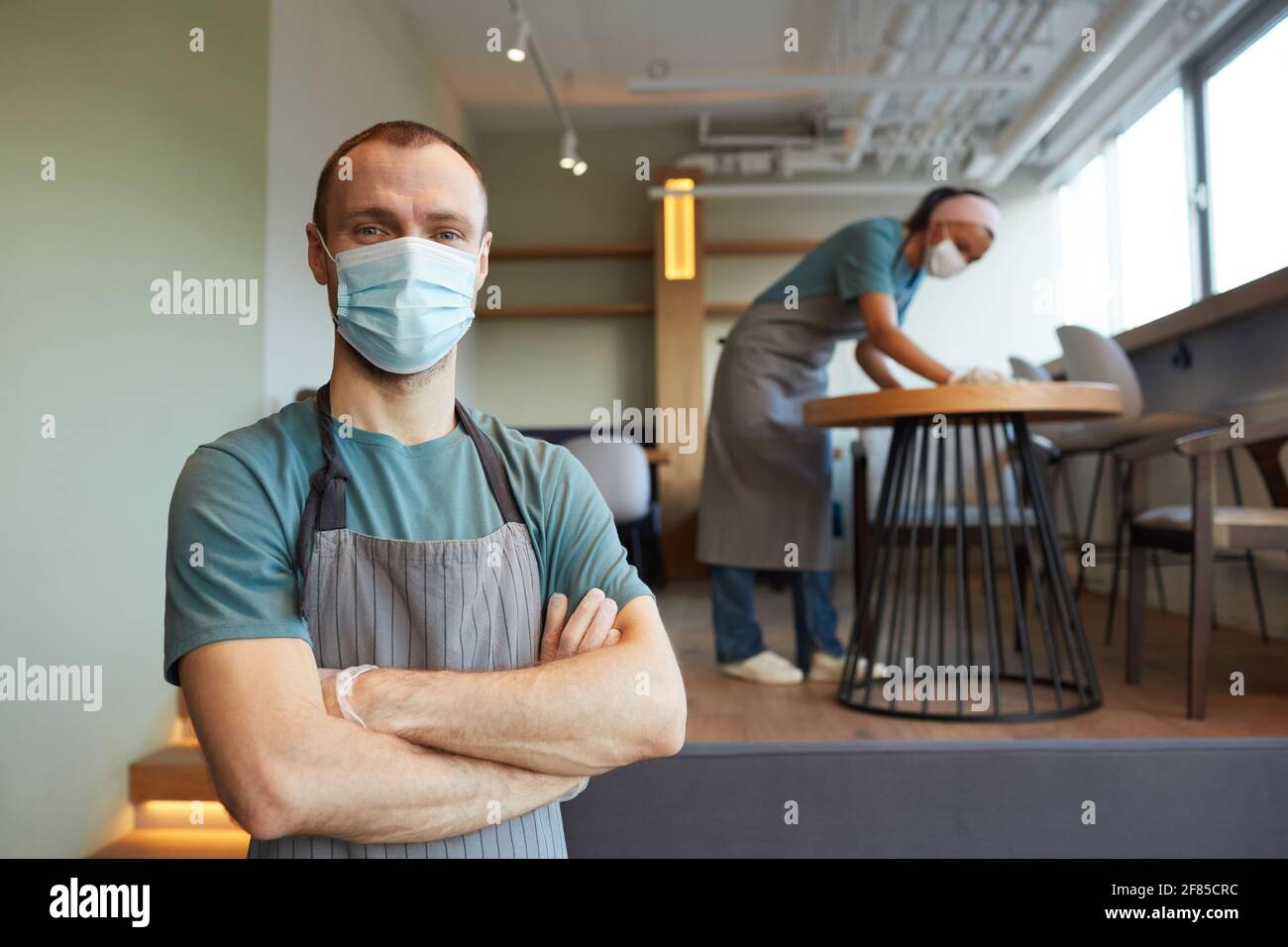Waist up portrait of young male waiter wearing mask and apron looking at camera while standing in cafe with woman cleaning in background, covid safety Stock Photo