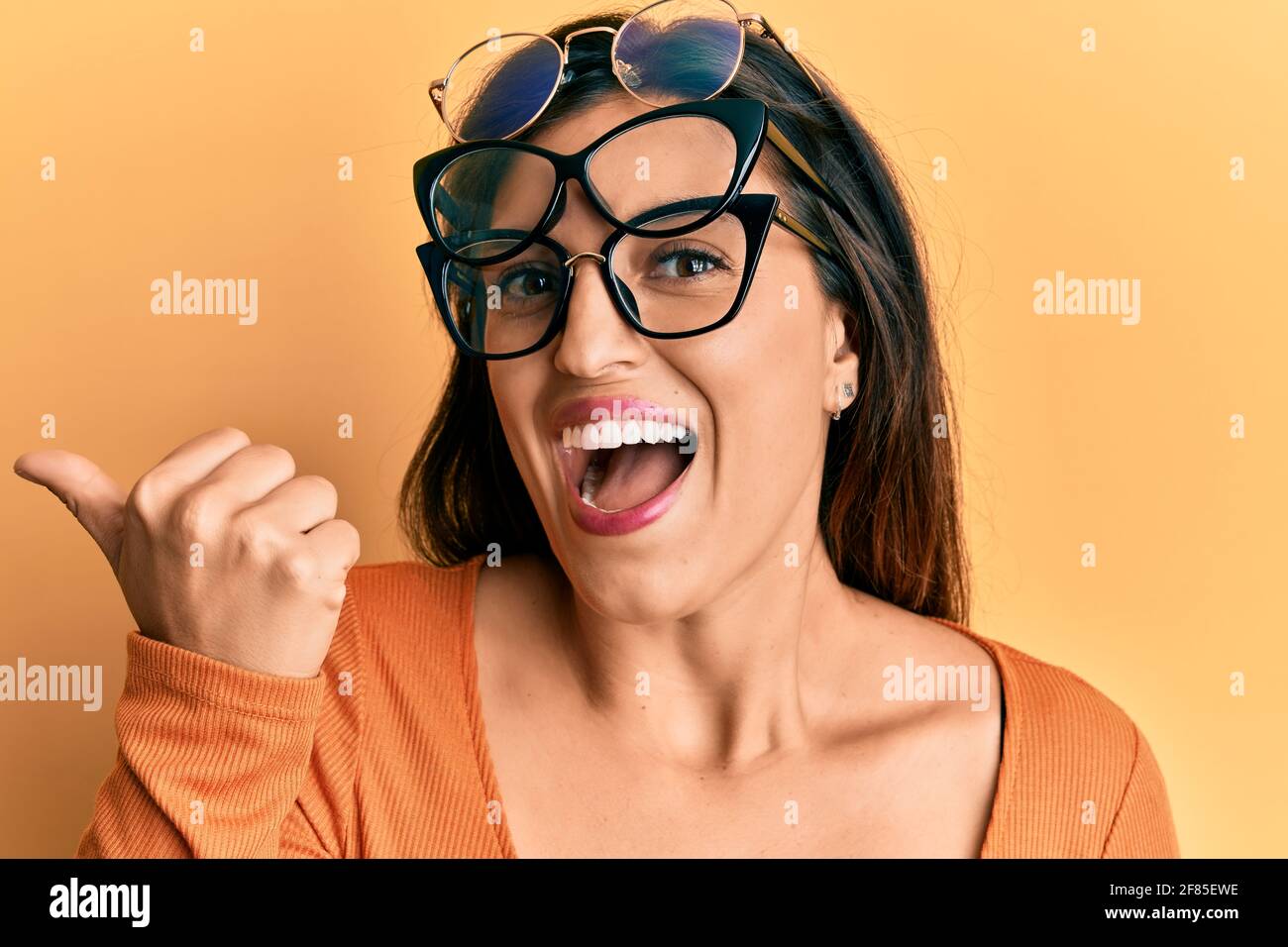 Beautiful hispanic woman wearing many glass over face pointing thumb up to the side smiling
