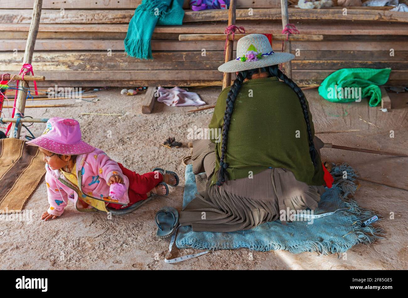 Bolivian indigenous Jalq'a woman weaving a textile with her daughter playing on the floor in Potolo, Sucre department, Bolivia. Stock Photo