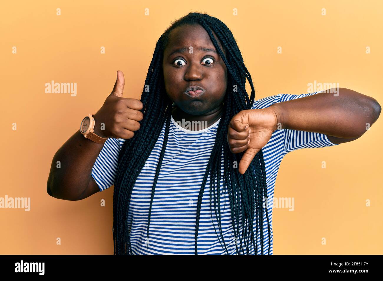Young black woman with braids doing thumbs down and thumbs up gesture puffing cheeks with funny face. mouth inflated with air, catching air. Stock Photo