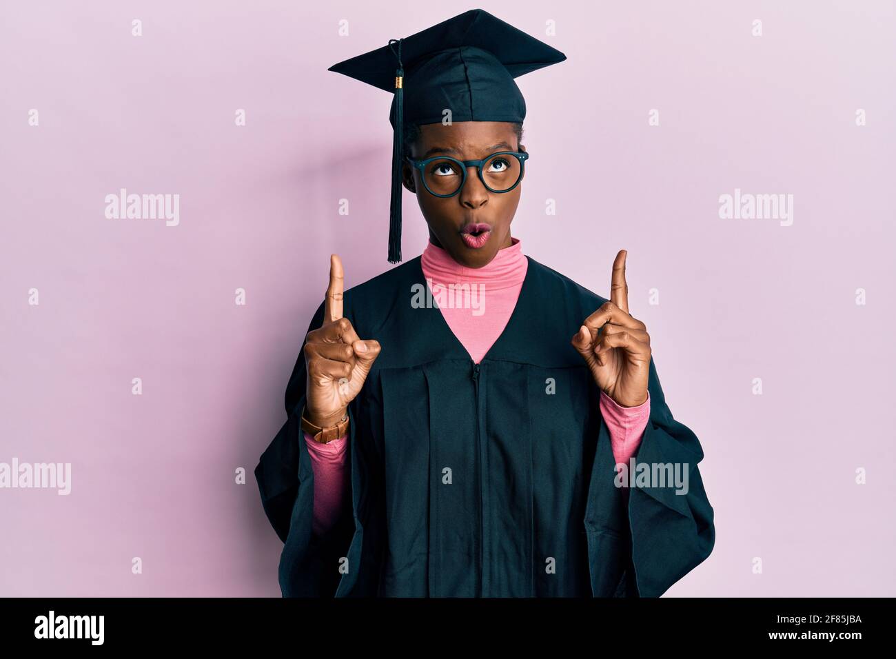 Young african american girl wearing graduation cap and ceremony robe amazed and surprised looking up and pointing with fingers and raised arms. Stock Photo