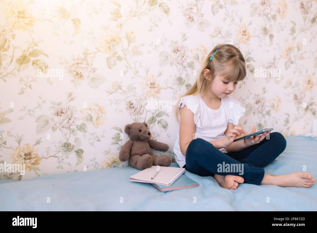 A little girl sits on the bed in the stylish bedroom, holding phone and reads something in smartphone. Communication concept Stock Photo