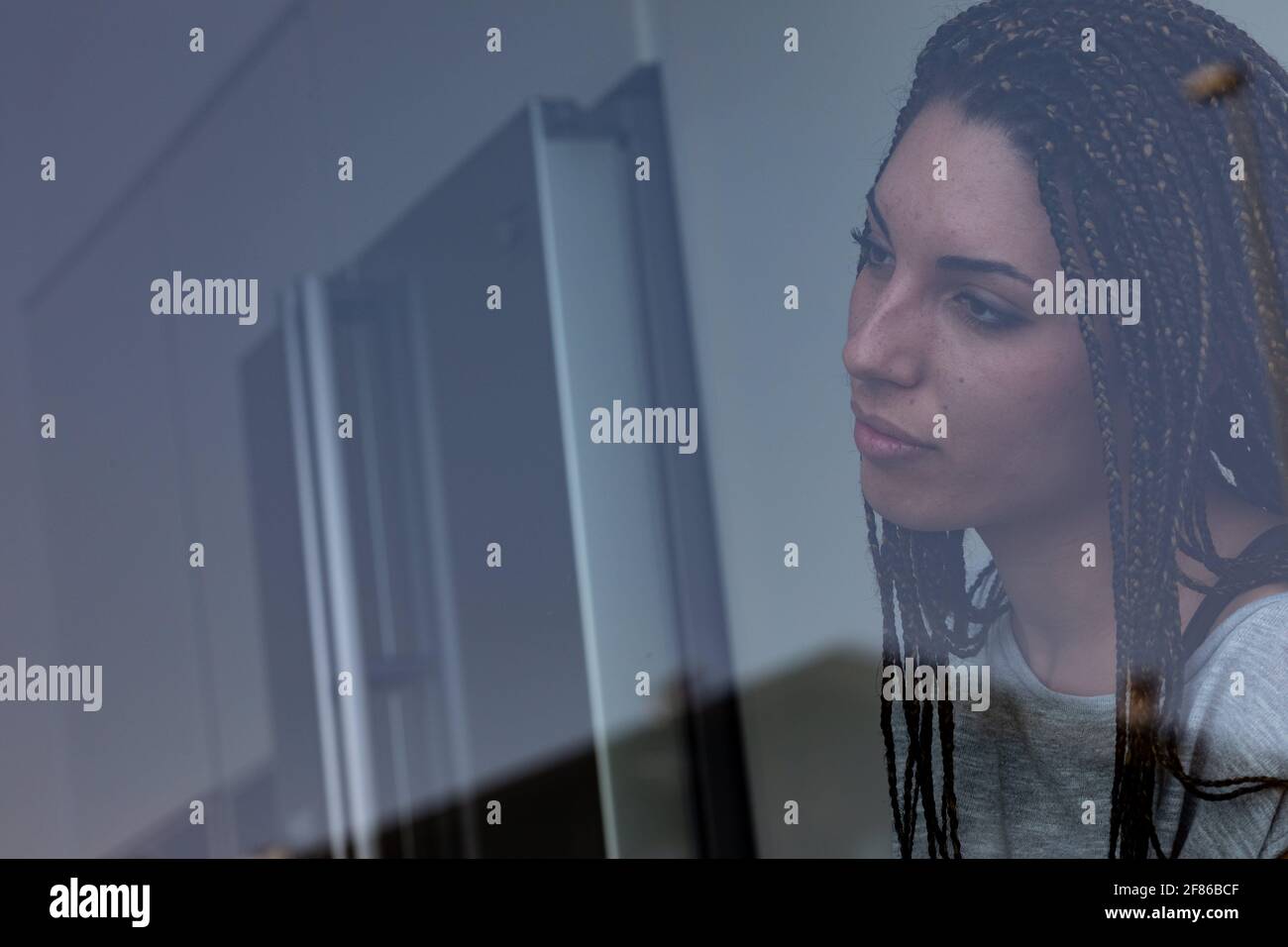 Lonely young woman with her hair braided in dreadlocks staring out of a window with look of longing and head tilted to the side with lateral copyspace Stock Photo
