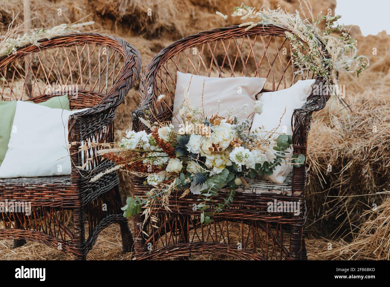 Two wicker chairs with white cushions and a bouquet of flowers on it. Rattan chairs with straw bales in the background. Boho style Stock Photo