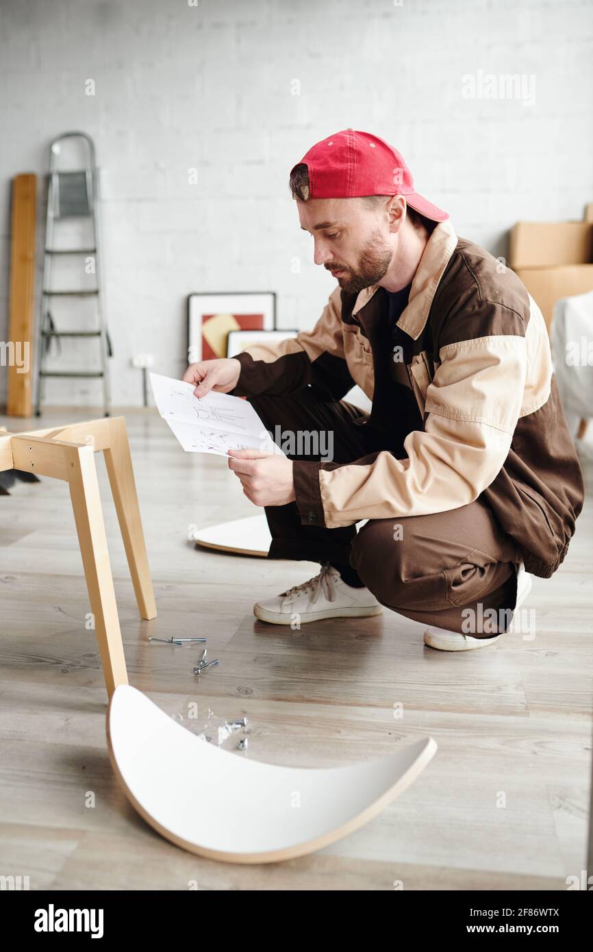 Serious assemblage specialist in workwear standing on squats and looking through instruction on paper while assembling furniture of client Stock Photo