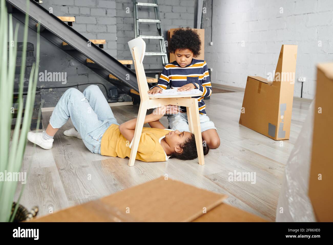 Cute mixed-race boy helping his sister with assembling wooden chair while standing on knees on the floor of living-room in their new flat Stock Photo