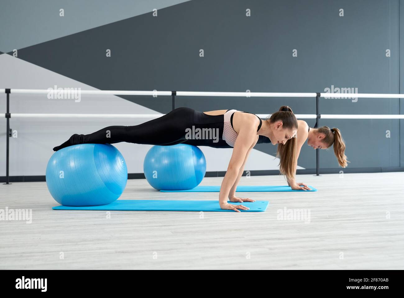 Side view of two attractive fit women training core muscles with legs on big blue fitness balls in studio with handrails. Gorgeous fitnesswomen practicing plank position on mats. Concept of sport. Stock Photo