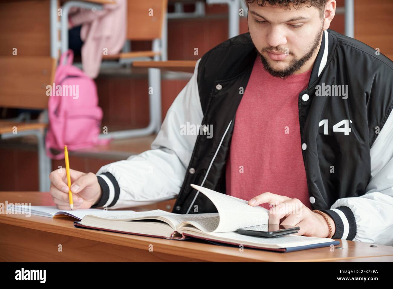 Young male student cheating while sitting by desk, making notes in copybook and looking at prompt on smartphone screen at lesson Stock Photo