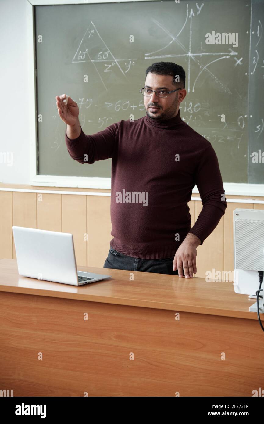 Mixed-race male in casualwear holding piece of chalk while standing between desk and blackboard and explaining something to students Stock Photo